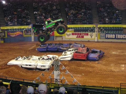 1996 Ford Bronco - A green monster truck soars through the air over a pile of destroyed cars during a Monster Jam event.