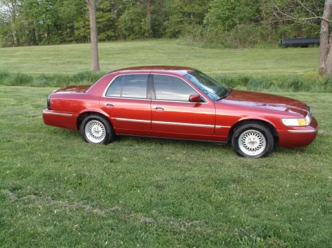 1999 Mercury Grand Marquis - This red 1999 Mercury Grand Marquis is parked on a grassy area with trees in the background.