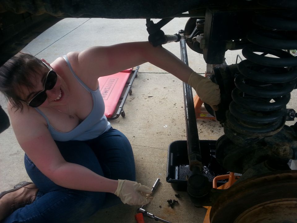 Woman in sunglasses and jeans works on vehicle suspension with tools nearby. A red creeper is behind her