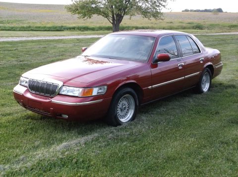 1999 Mercury Grand Marquis - A red sedan with a flat tire sits on a grassy field under a tree.