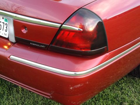 1999 Mercury Grand Marquis - Rear view of a red Mercury sedan showing its taillight and chrome trim, with visible scratches on the bumper.