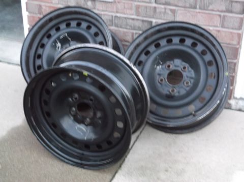 1999 Mercury Grand Marquis - Four black steel wheels are stacked on a concrete surface in front of a brick wall.
