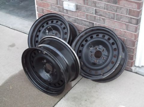 1999 Mercury Grand Marquis - A set of four black steel wheels are shown resting on a concrete surface next to a brick wall.