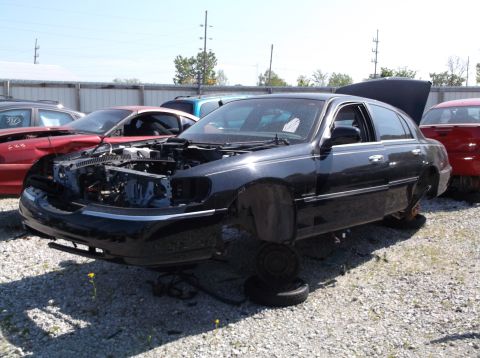 1999 Mercury Grand Marquis - Junkyard scene with a black sedan missing many parts, including wheels and engine components.