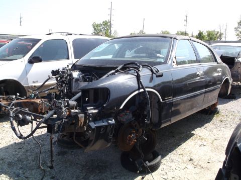 1999 Mercury Grand Marquis - A dark sedan sits in a junkyard with its front end completely dismantled, exposing the chassis and wiring.