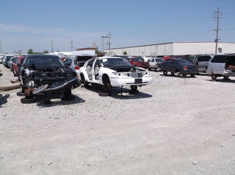 1999 Mercury Grand Marquis - Junkyard scene with a 1999 Mercury Grand Marquis and other vehicles, all missing parts.