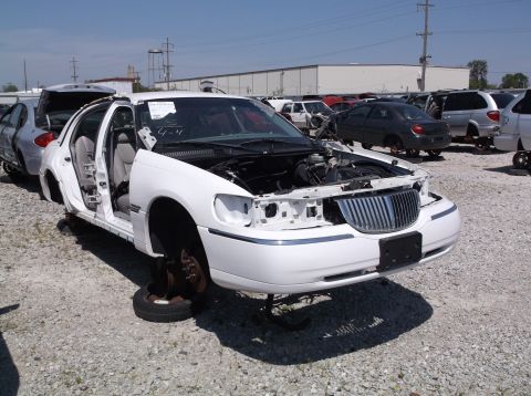 1999 Mercury Grand Marquis - This white sedan is missing its doors and wheels, with its hood open, in a junkyard setting.