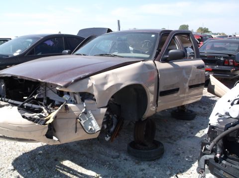 1999 Mercury Grand Marquis - A damaged sedan sits in a junkyard, missing its wheels and showing significant front-end collision damage.
