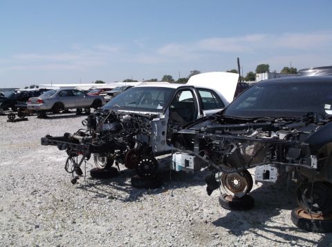 1999 Mercury Grand Marquis - Two stripped car chassis sit in a junkyard under a clear blue sky.