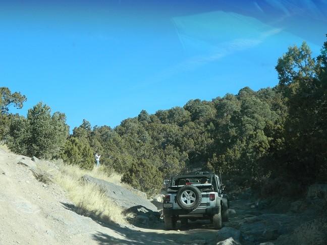 Silver Jeep Wrangler with spare tire on back driving on a rocky dirt road with trees and blue sky