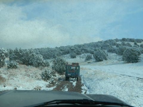 1979 Ford Bronco - Two vehicles travel on a snowy dirt road, surrounded by snow-dusted trees under a cloudy sky.