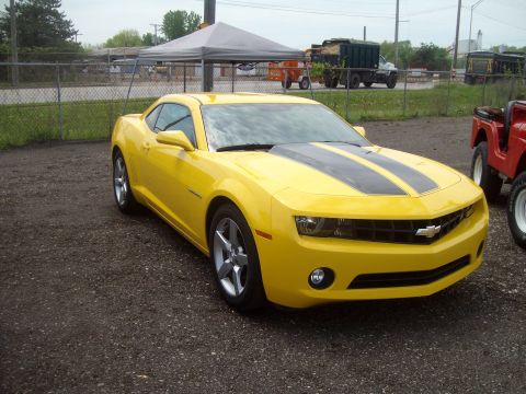 2004 Ford Crown Victoria - A yellow Chevrolet Camaro with black racing stripes is parked on a gravel lot next to a red Jeep.