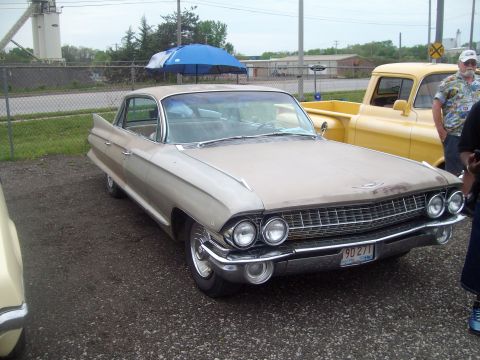 2004 Ford Crown Victoria - A classic Cadillac with prominent fins sits parked on a gravel lot next to a yellow pickup truck.