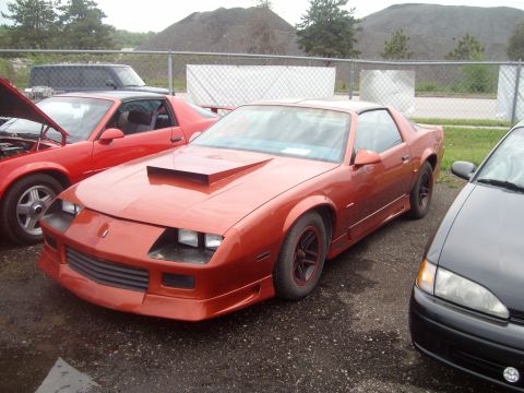 2004 Ford Crown Victoria - An orange third-generation Camaro is parked next to a red Camaro with its hood open.