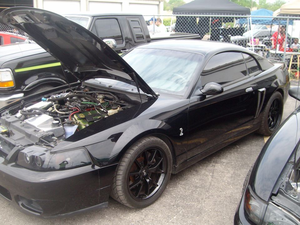 Black Ford Mustang coupe with hood open showing engine bay. Another vehicle is parked to the left, and part of a third vehicle is visible on the right