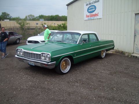 2004 Ford Crown Victoria - A green 1964 Chevrolet Impala sedan with a white roof and gold wire wheels is parked outside an auto body shop.