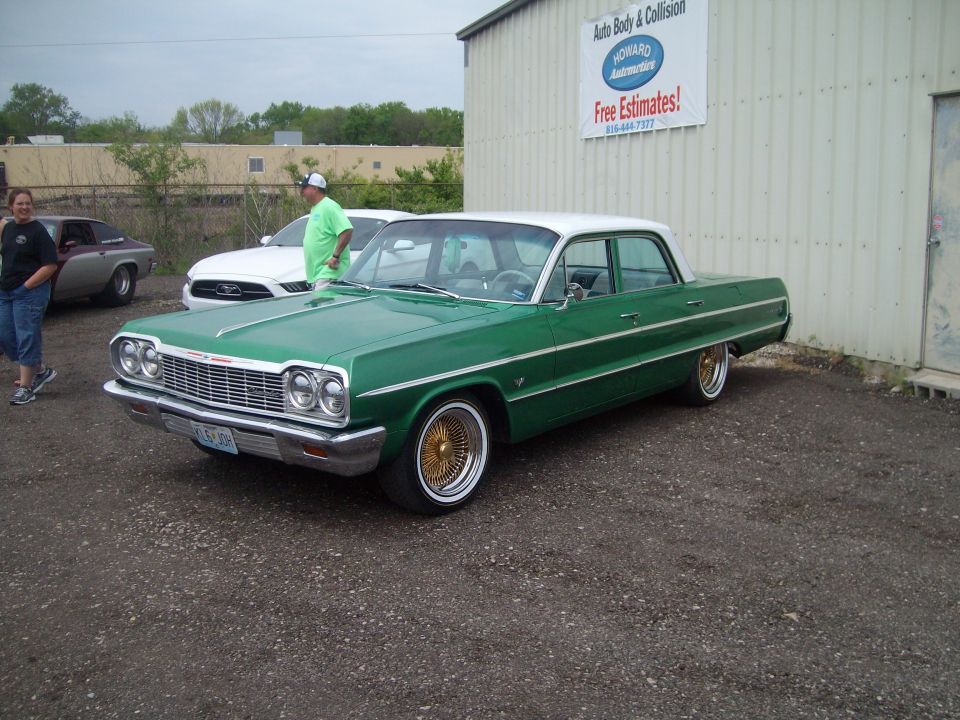 Green 1964 Chevrolet Impala sedan with a white roof and gold wire wheels parked outside a garage