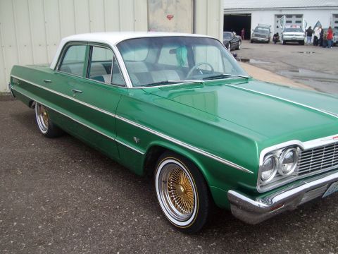 2004 Ford Crown Victoria - A green and white 1964 Chevrolet Impala sedan is parked outside a building, with other vehicles visible in the background.