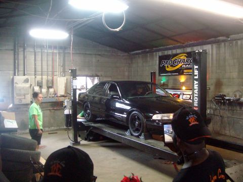 2004 Ford Crown Victoria - A black sedan is positioned on a rotary lift inside a garage, with onlookers present.