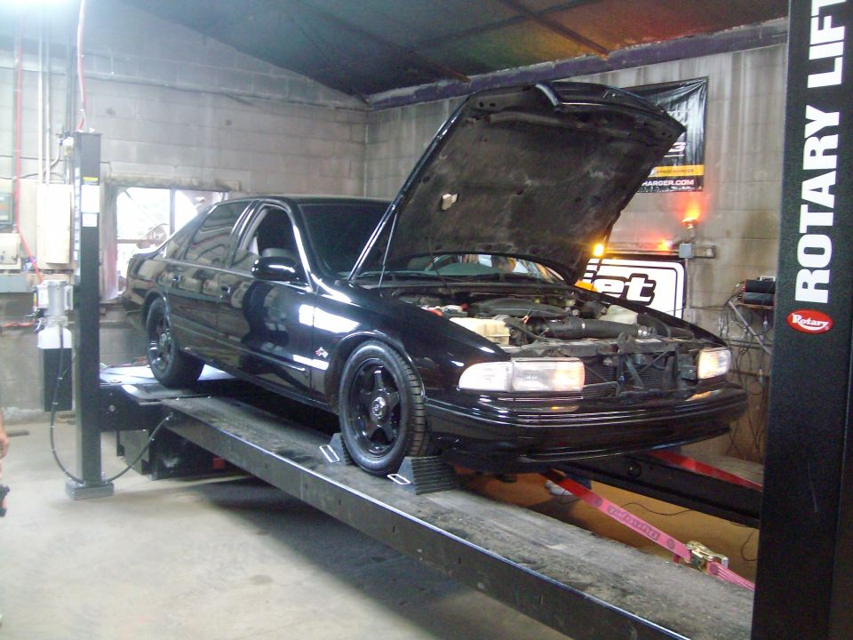 Black sedan with hood open on a car lift, front view. Aftermarket wheels and some engine components visible