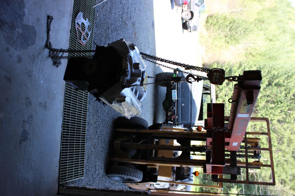 engine and transmission assembly suspended by a chain hoist over a forklift, with a dark SUV visible in the background
