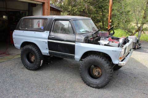 1979 Ford Bronco - Lifted two-tone gray and black Ford Bronco with mud-terrain tires and an exposed V8 in a shop driveway.