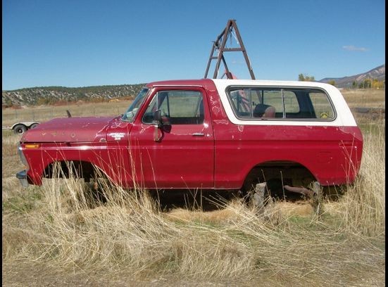 Red 1978 Ford Bronco with white top, missing wheels, parked in dry grass