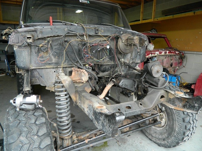 Disassembled truck chassis showing engine, suspension, and wiring. Another red vehicle is partially visible behind it