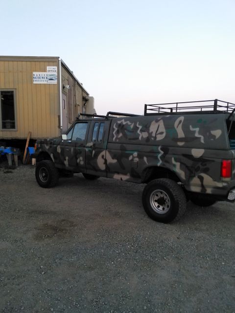 1991 Ford Bronco - A four-door pickup truck with a camouflage paint job and a camper shell is parked on a gravel lot.