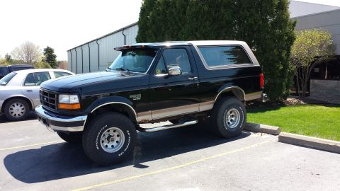1995 Ford Bronco - This lifted Ford Bronco is parked in a lot, with a silver sedan nearby and a building in the background.
