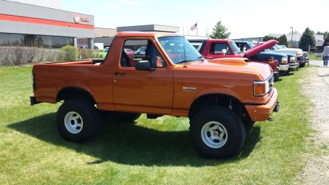 1995 Ford Bronco - A line of trucks and SUVs are parked on a grassy area outside a building, with the orange Bronco in the foreground.