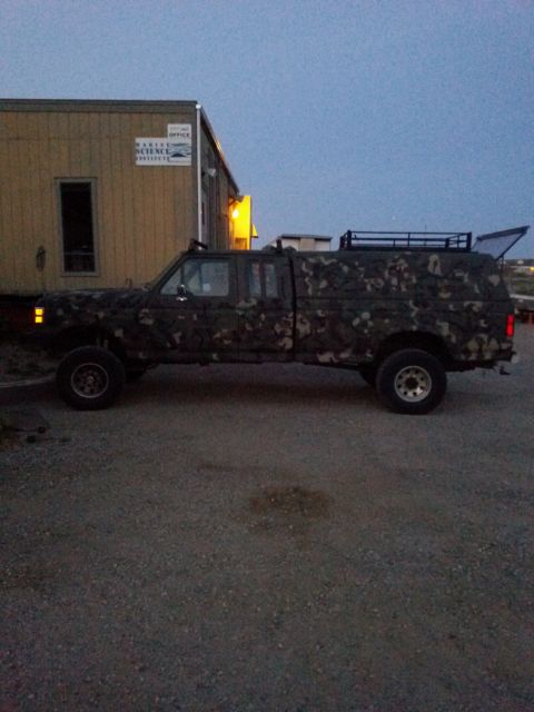 1991 Ford Bronco - A camouflaged pickup truck with a camper shell is parked in front of a building with a "Marine Science Institute" sign.