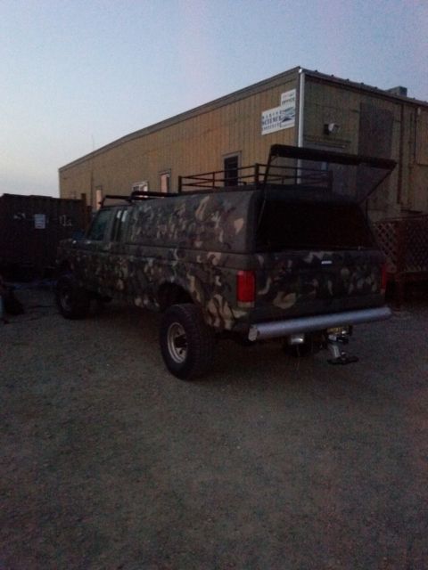 1991 Ford Bronco - This camouflaged Ford F-Series truck is equipped with a camper shell and roof rack, seen parked outdoors.