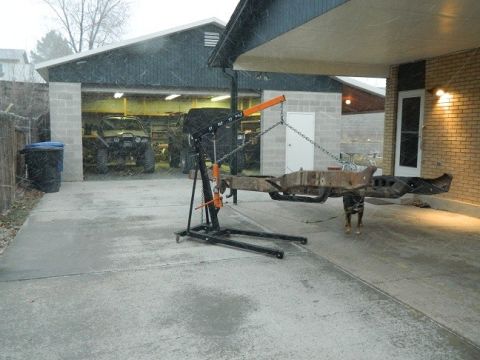 1979 Ford Bronco - A vehicle frame is suspended by a hoist in front of a garage where two SUVs are parked.