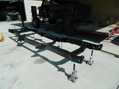 1979 Ford Bronco - A vehicle frame, freshly powder coated, sits on jack stands outside a garage. A dark SUV body is visible on a trailer behind it.