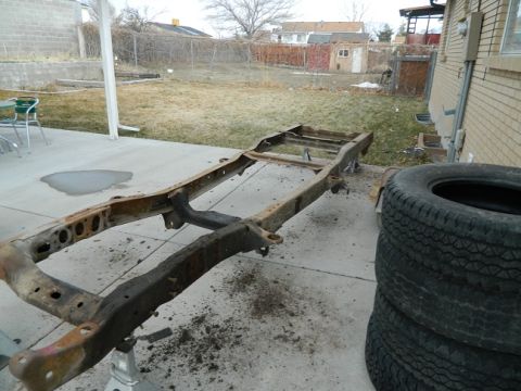 1979 Ford Bronco - A bare vehicle frame sits on jack stands in a backyard, with a stack of tires to the right.