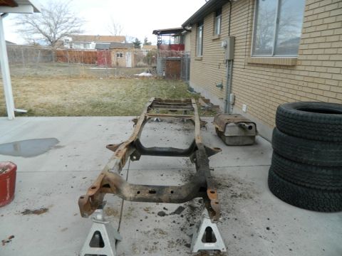 1979 Ford Bronco - A vehicle frame, likely from a Ford Bronco, sits on jack stands in a backyard, with a fuel tank and tires stacked to the side.