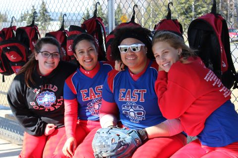 1990 Chevrolet Suburban - Four young women in East softball uniforms smile for a photo while sitting in front of a fence with backpacks hanging.