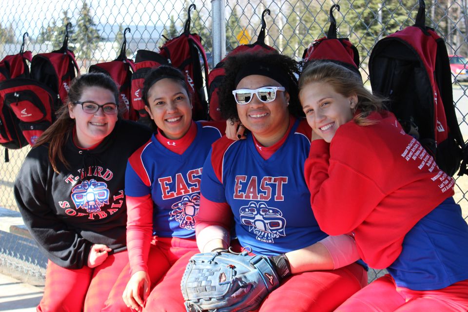 Four smiling young women in softball uniforms pose for a photo