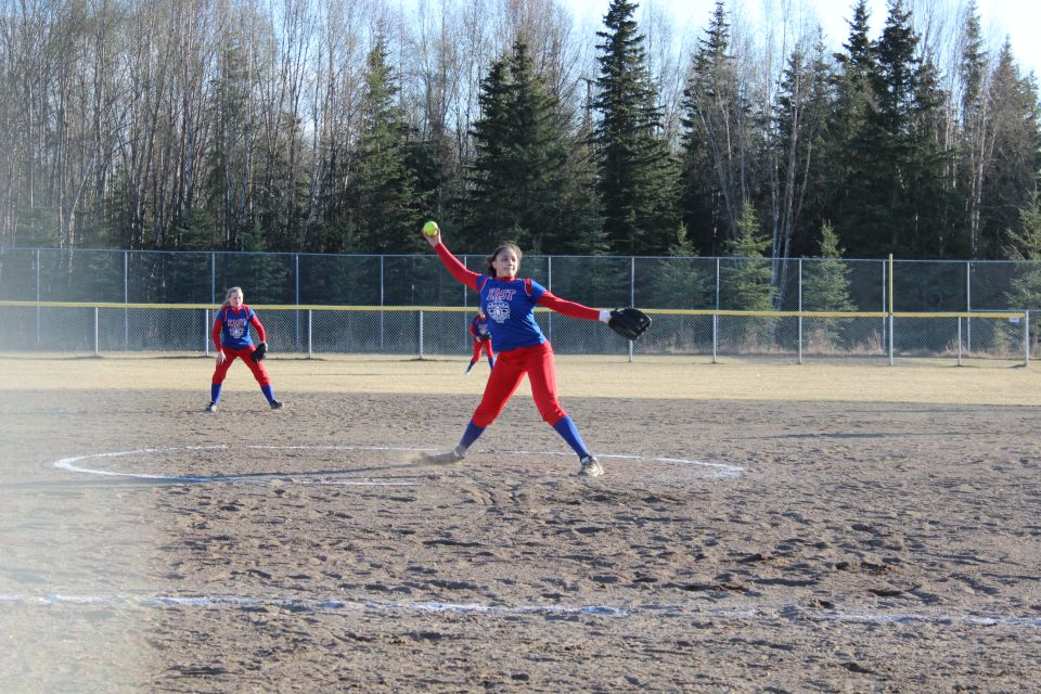 Softball pitcher in red pants and blue shirt winds up to throw on a dirt field