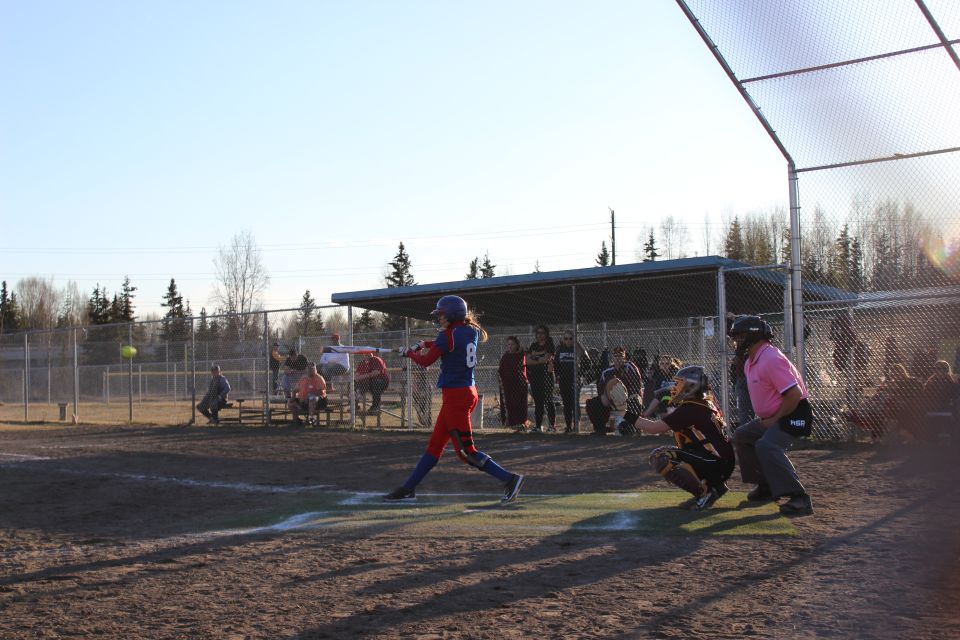 Batter swings at a pitch during a softball game, with a catcher and umpire behind home plate. Spectators watch from the bleachers