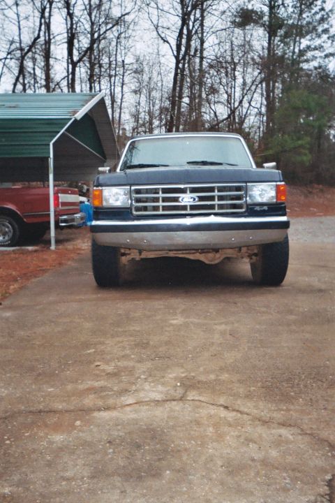 1988 Ford Bronco - A lifted dark blue Ford truck is parked in front of a carport, with another vehicle visible to the left.
