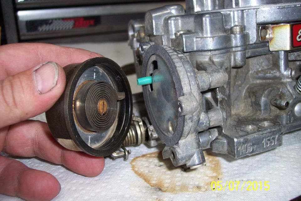 Hand holding a disassembled carburetor spring and plunger assembly next to the main carburetor body. Part number 1406 is visible