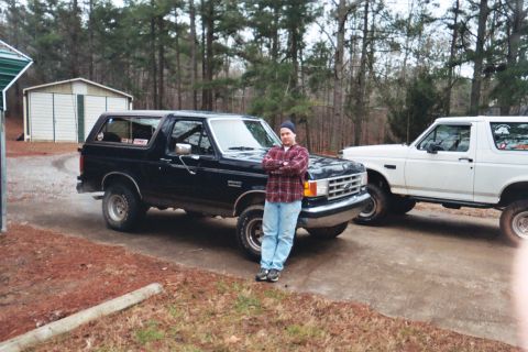1988 Ford Bronco - A man poses in front of a lifted black 1988 Ford Bronco, with a white Bronco visible behind it.
