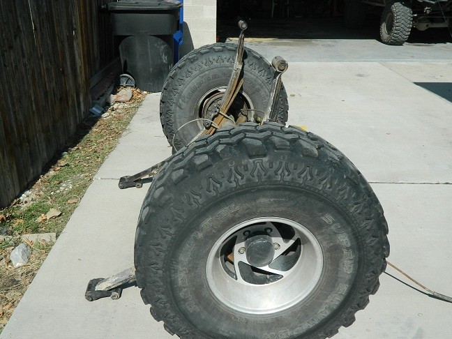 Rear axle and large off-road tires on a concrete surface, with another vehicle in the background
