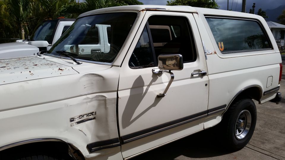 White Ford Bronco with damage to the front fender. Another white vehicle is parked behind it
