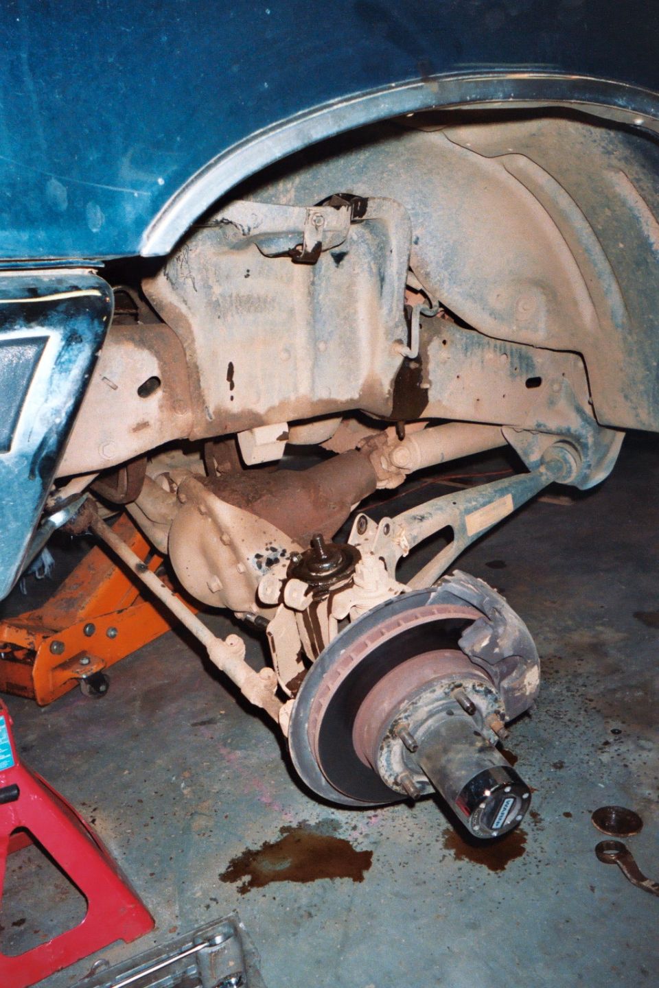 close-up of a vehicle's front wheel hub, brake rotor, and suspension components, with a jack stand and floor jack visible