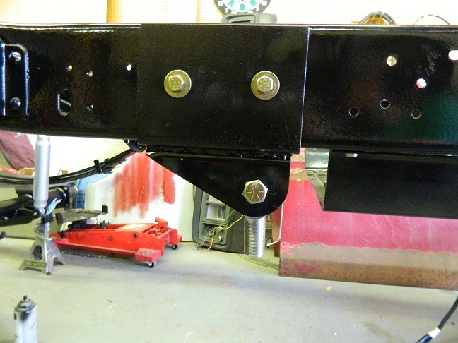 Close-up of black painted vehicle frame with suspension bracket and bolts. Red floor jack and jack stands visible below