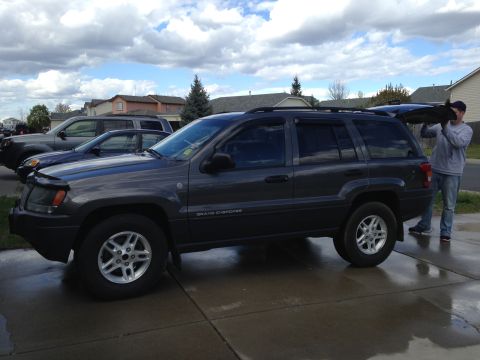 2004 Jeep Grand Cherokee - A man loads an item into the back of a parked Jeep Grand Cherokee, with other vehicles visible in the background.