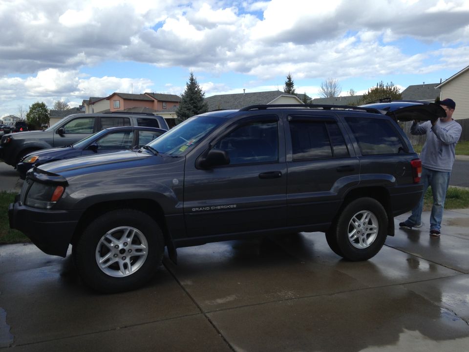 Dark gray Jeep Grand Cherokee parked in driveway, another SUV and truck behind it. Man is putting something in the rear cargo area